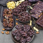 Various chocolate-covered nuts, including peanuts and almonds, arranged in bowls and trays on a dark surface. Whole almonds and peanuts scattered around.