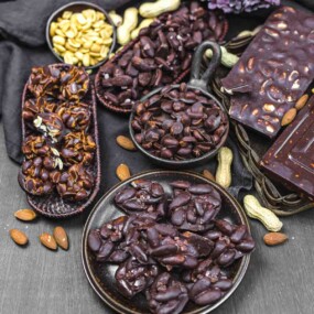 Various chocolate-covered nuts, including peanuts and almonds, arranged in bowls and trays on a dark surface. Whole almonds and peanuts scattered around.
