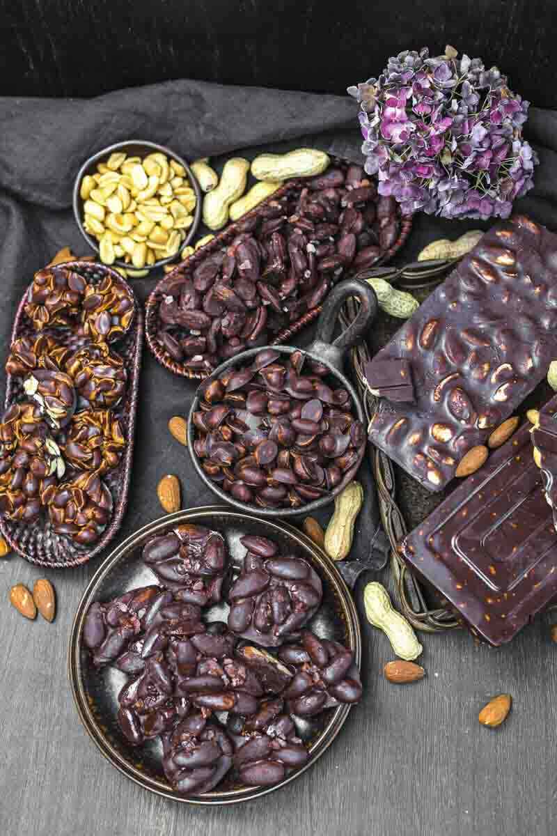 An assortment of dark chocolates with nuts, dried fruits, and whole peanuts arranged on a black surface next to a dark purple hydrangea flower.