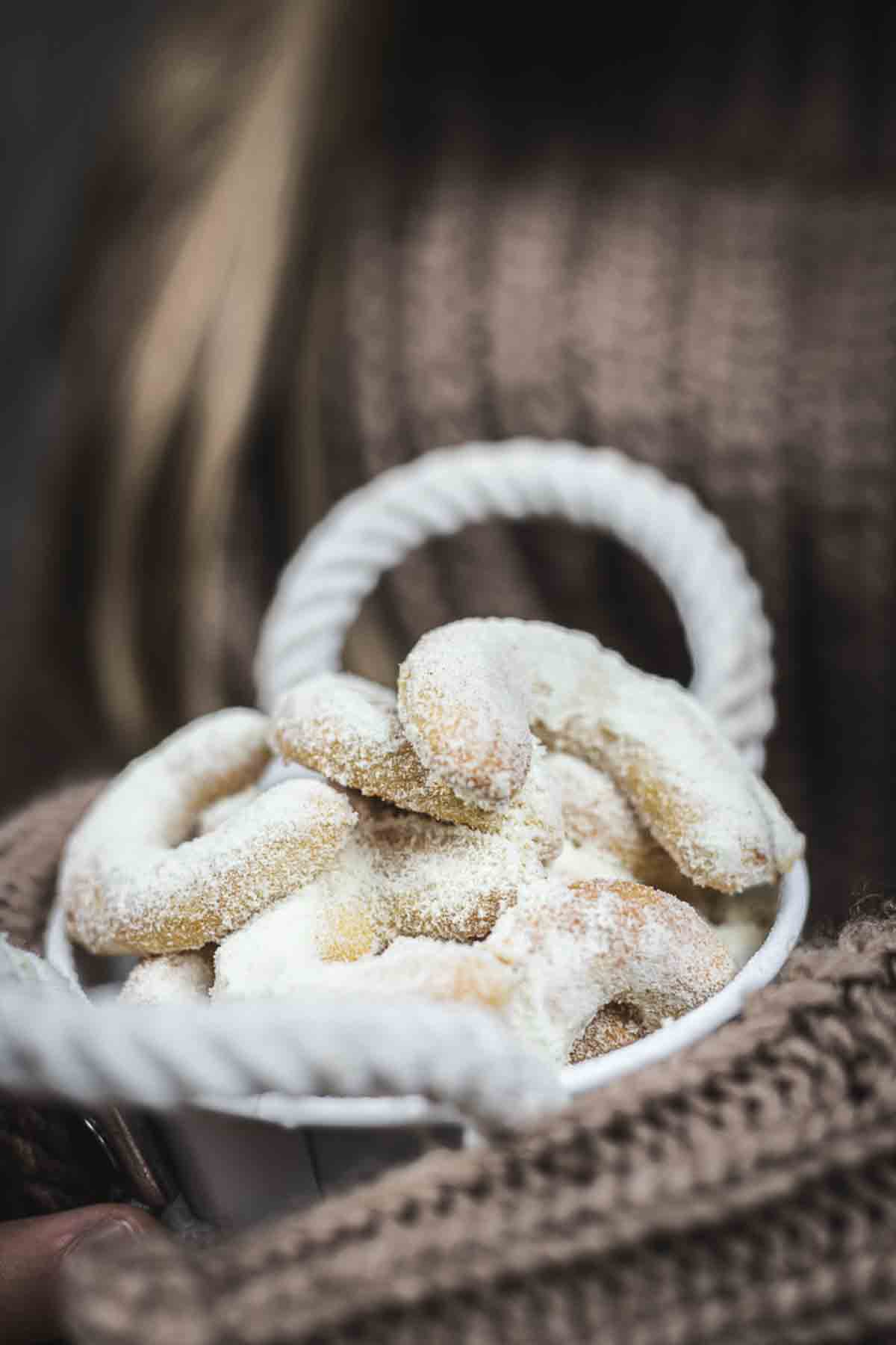 A white bowl filled with powdered sugar-covered crescent cookies, held by a person wearing a brown knitted sweater.