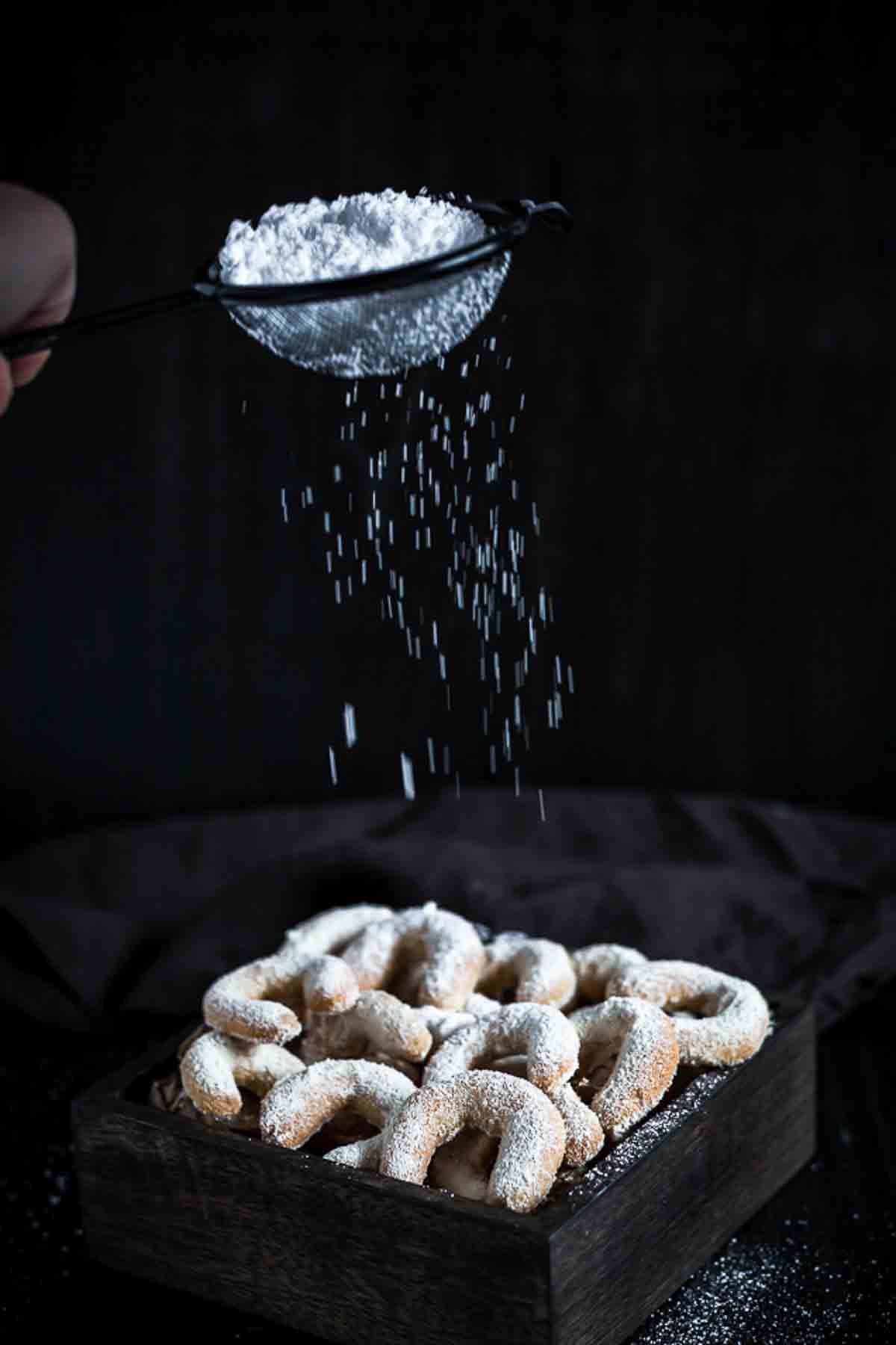 Powdered sugar is being sifted over a tray of crescent-shaped cookies on a dark background.