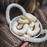 A bowl of crescent-shaped cookies dusted with powdered sugar, held by a person wearing a knitted sweater.
