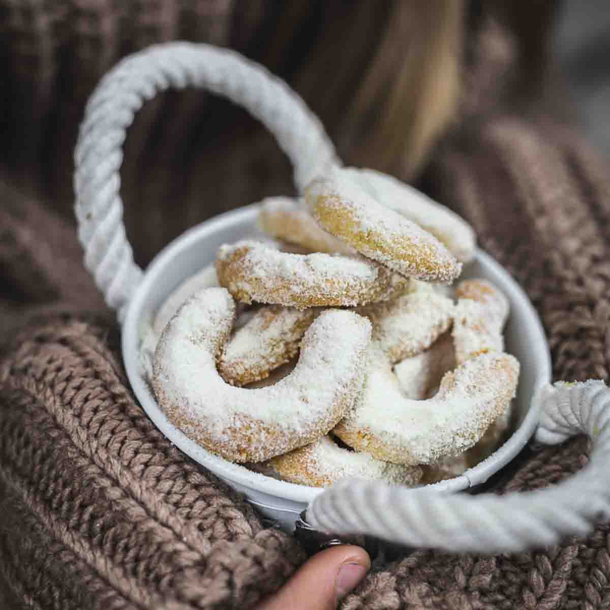 A bowl of crescent-shaped cookies dusted with powdered sugar, held by a person wearing a knitted sweater.