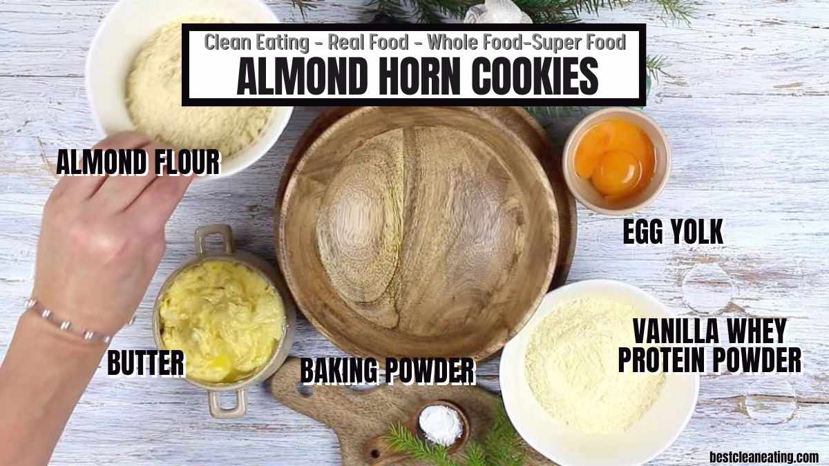 Ingredients for almond horn cookies arranged on a table: almond flour, butter, baking powder, egg yolk, and vanilla whey protein powder. An empty wooden bowl is in the center.