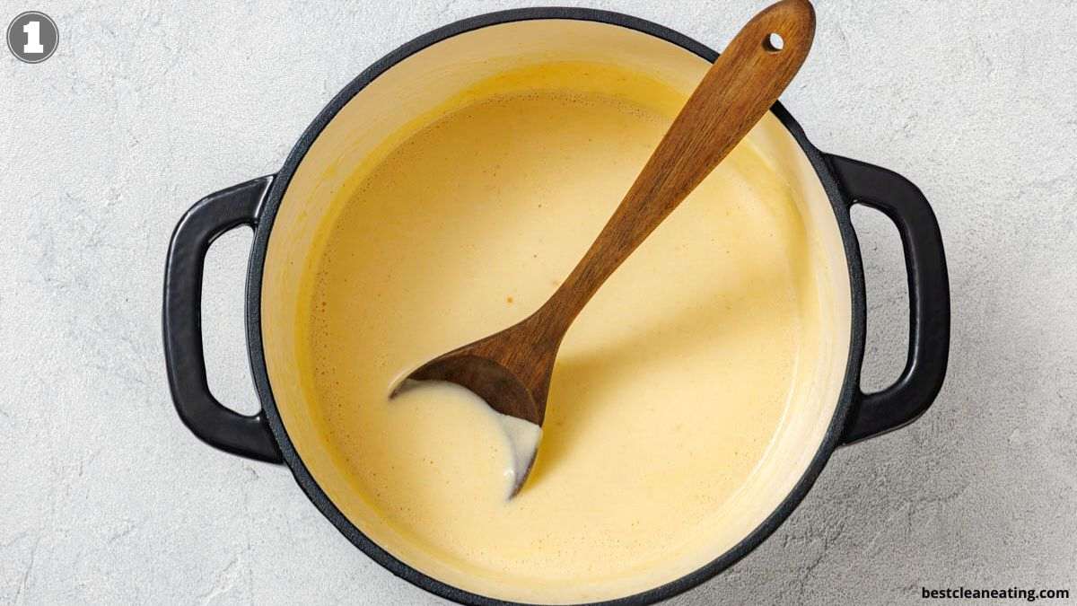 A black pot with creamy yellow soup or sauce inside, being stirred with a wooden spoon, on a light-colored surface.