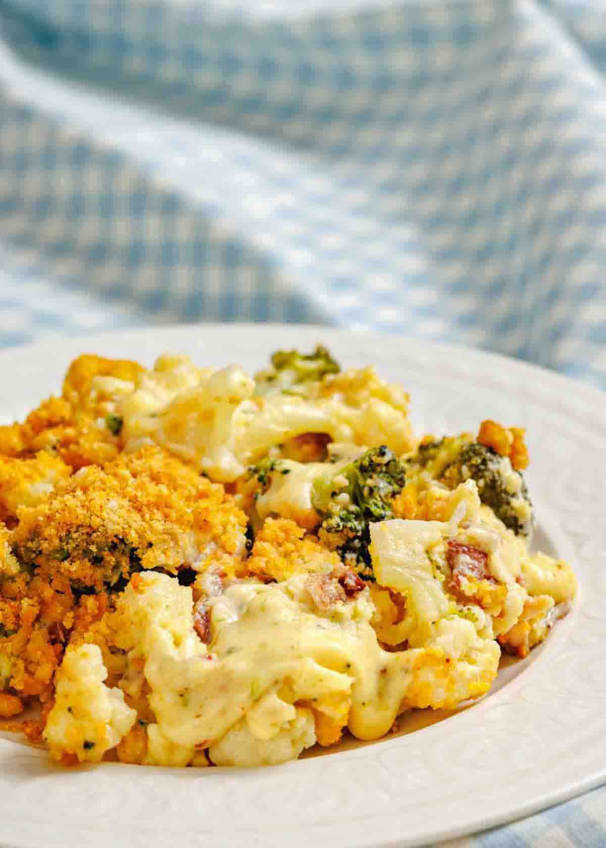 A plate of cheesy broccoli and cauliflower casserole topped with a golden breadcrumb crust, set against a blue and white checkered fabric background.
