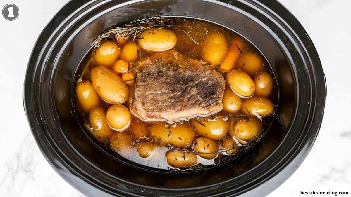 A slow cooker filled with a cooked beef roast, baby potatoes, carrots, and broth.