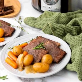 A plate of sliced pot roast with baby potatoes, carrots, and onions, garnished with rosemary, sits on a marble countertop near a slow cooker and green kitchen towel.