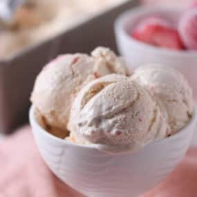 A white bowl filled with three scoops of strawberry ice cream sits on a light pink surface, with a bowl of strawberries and a container of ice cream in the background.