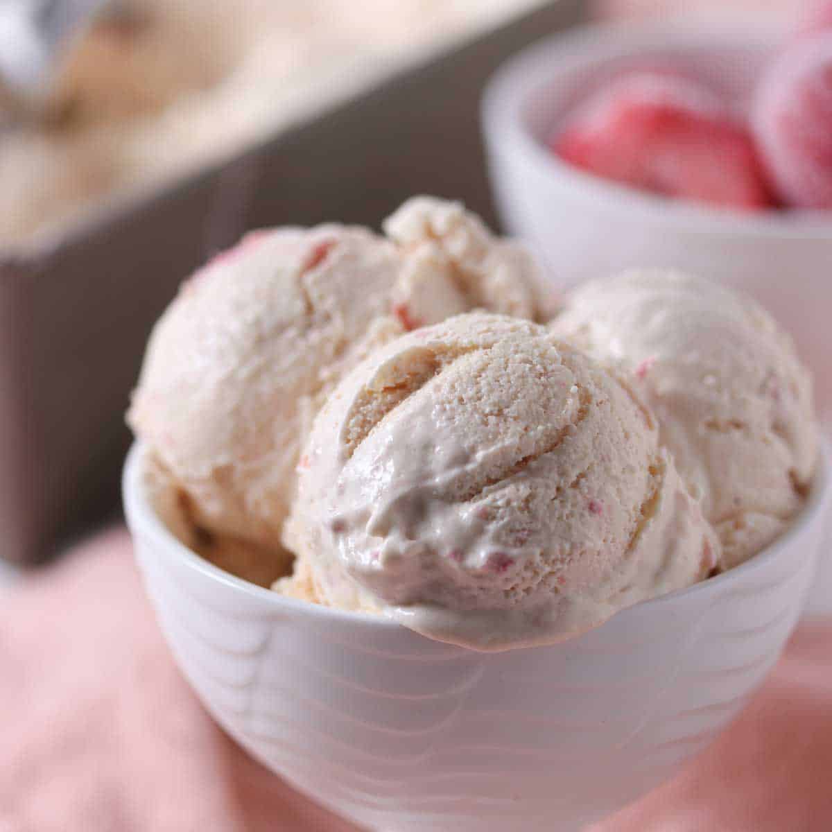 A white bowl filled with three scoops of strawberry ice cream sits on a light pink surface, with a bowl of strawberries and a container of ice cream in the background.