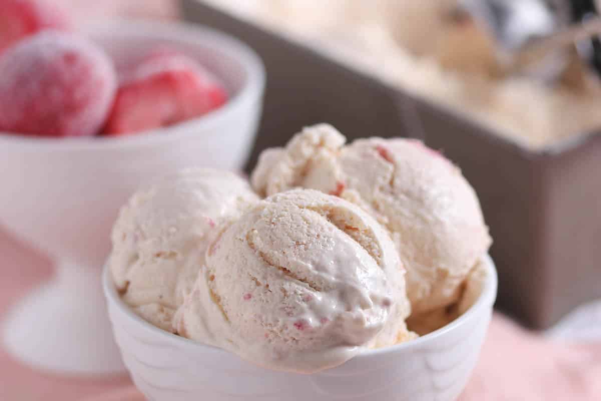 Three scoops of strawberry ice cream in a white bowl, with a bowl of frozen strawberries and an ice cream container in the background.