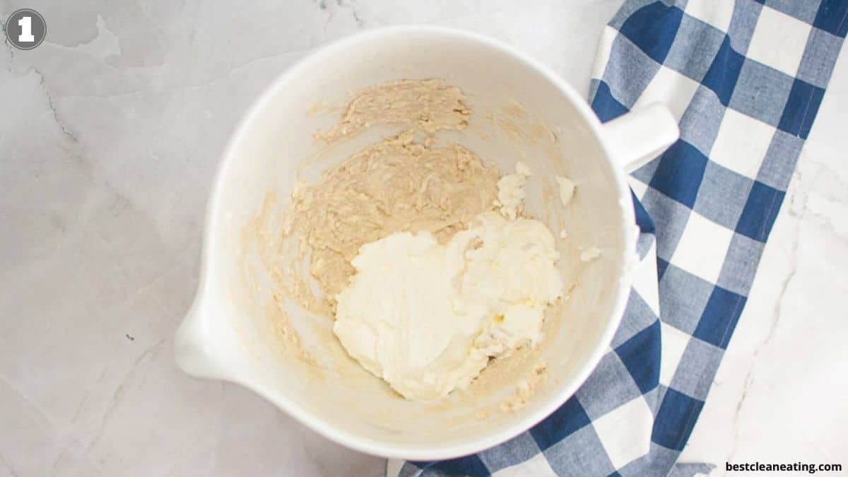 A white mixing bowl containing partially mixed batter and cream cheese on a marble surface with a blue plaid cloth.