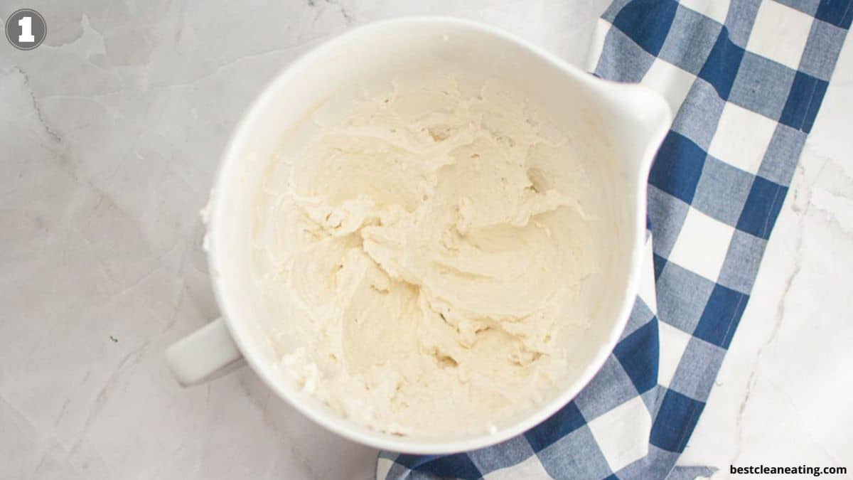 A white mixing bowl filled with creamy white batter sits on a marble surface next to a blue and white checkered cloth.