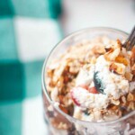 A close-up of a spoonful of parfait with whipped cream, berries, and granola in a glass, with a teal and white checkered cloth in the background.