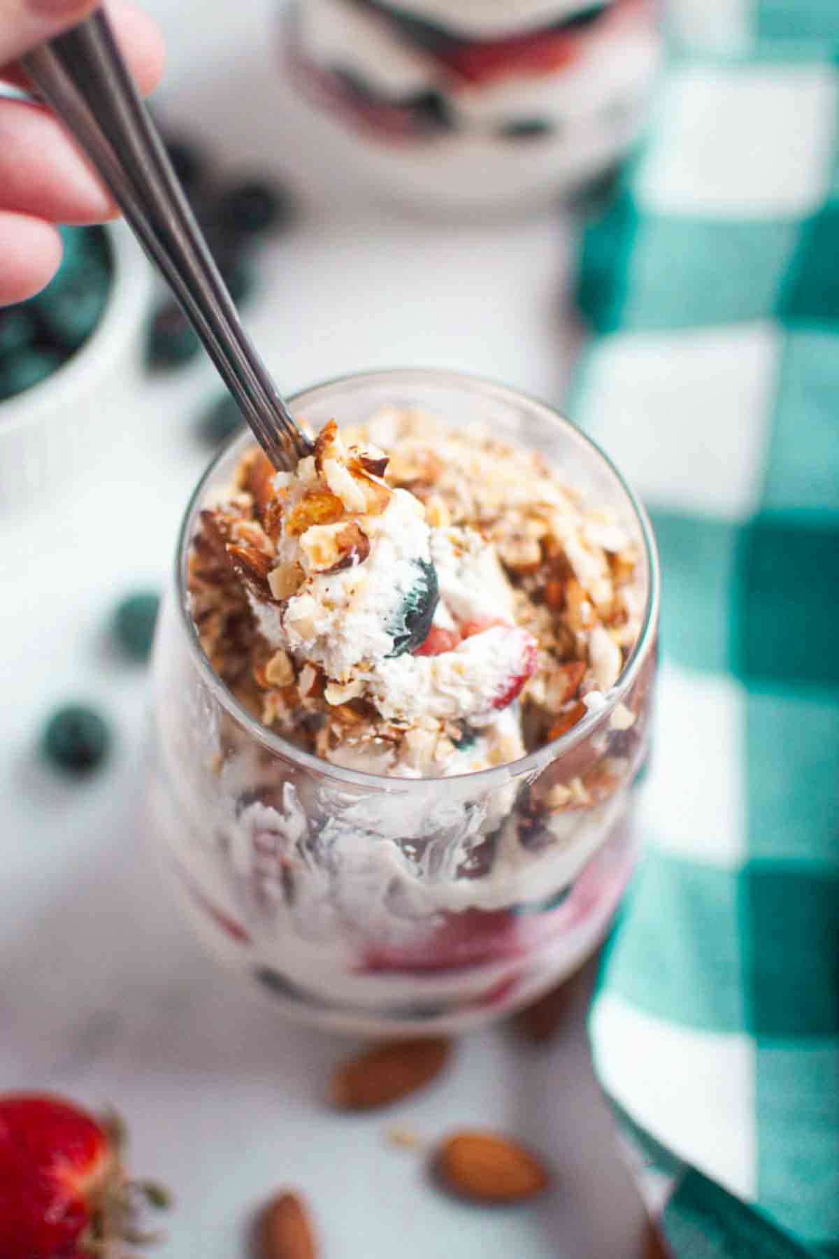 A hand holding a spoon prepares to eat a glass filled with yogurt, granola, and fruit, with whole almonds and a green plaid napkin nearby.