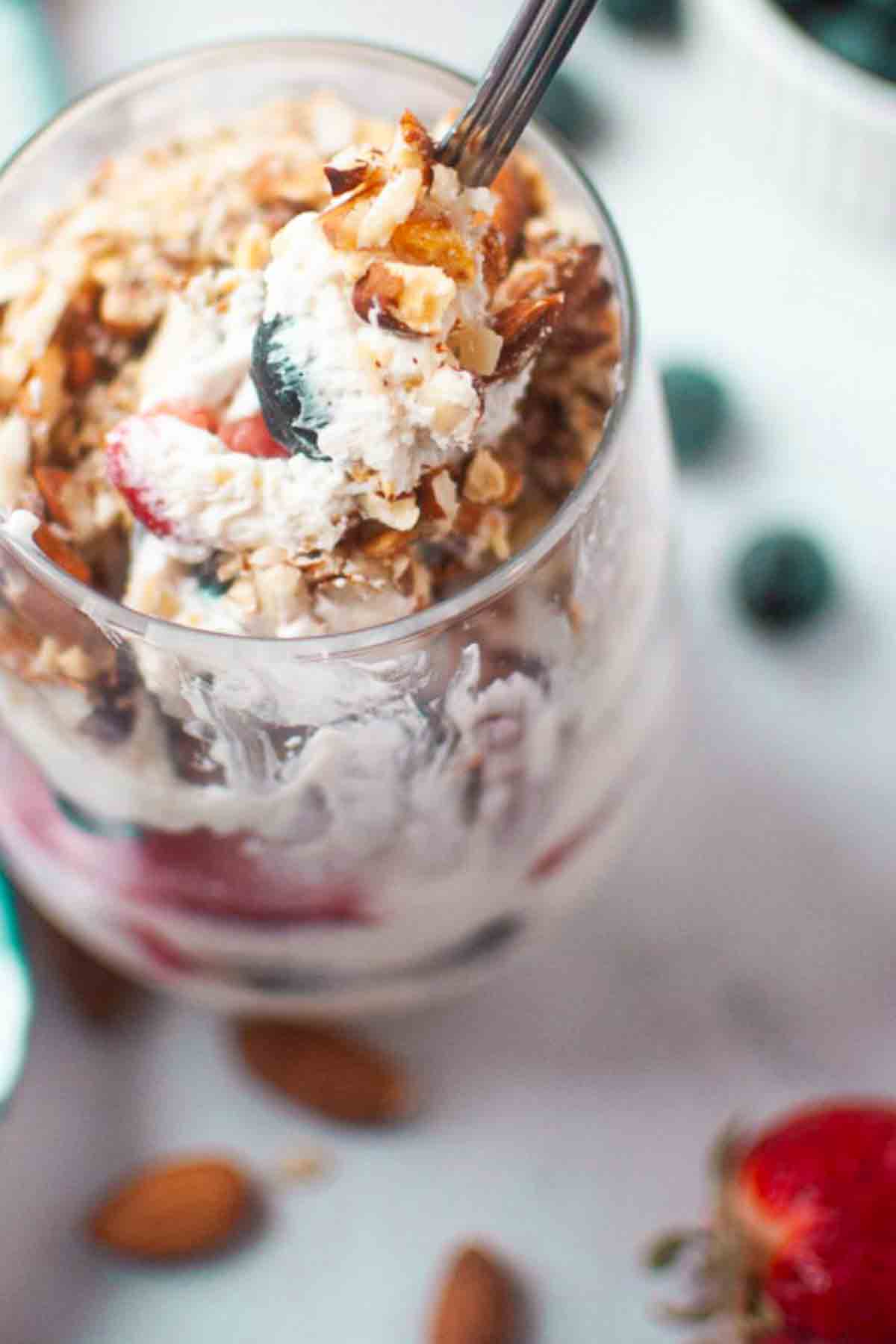 A glass filled with layers of whipped cream, berries, and granola, topped with chopped nuts, with a spoon scooping out a portion.