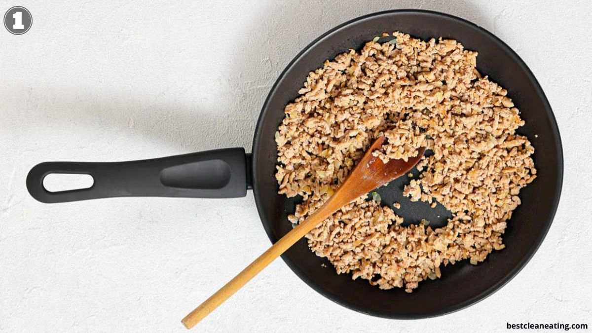 A nonstick frying pan with cooked ground meat being stirred with a wooden spoon on a white surface.