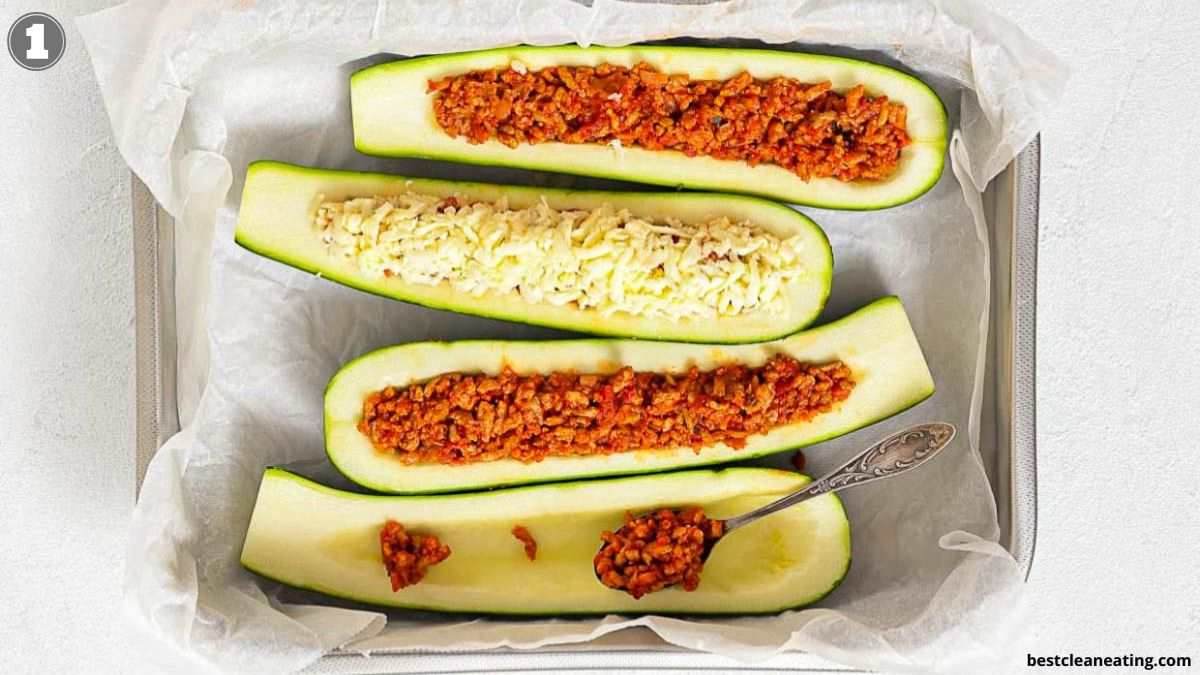 Four zucchini halves in a baking dish; three are filled with a tomato-based meat mixture, one with cheese and meat filling. A spoon rests in one zucchini half.