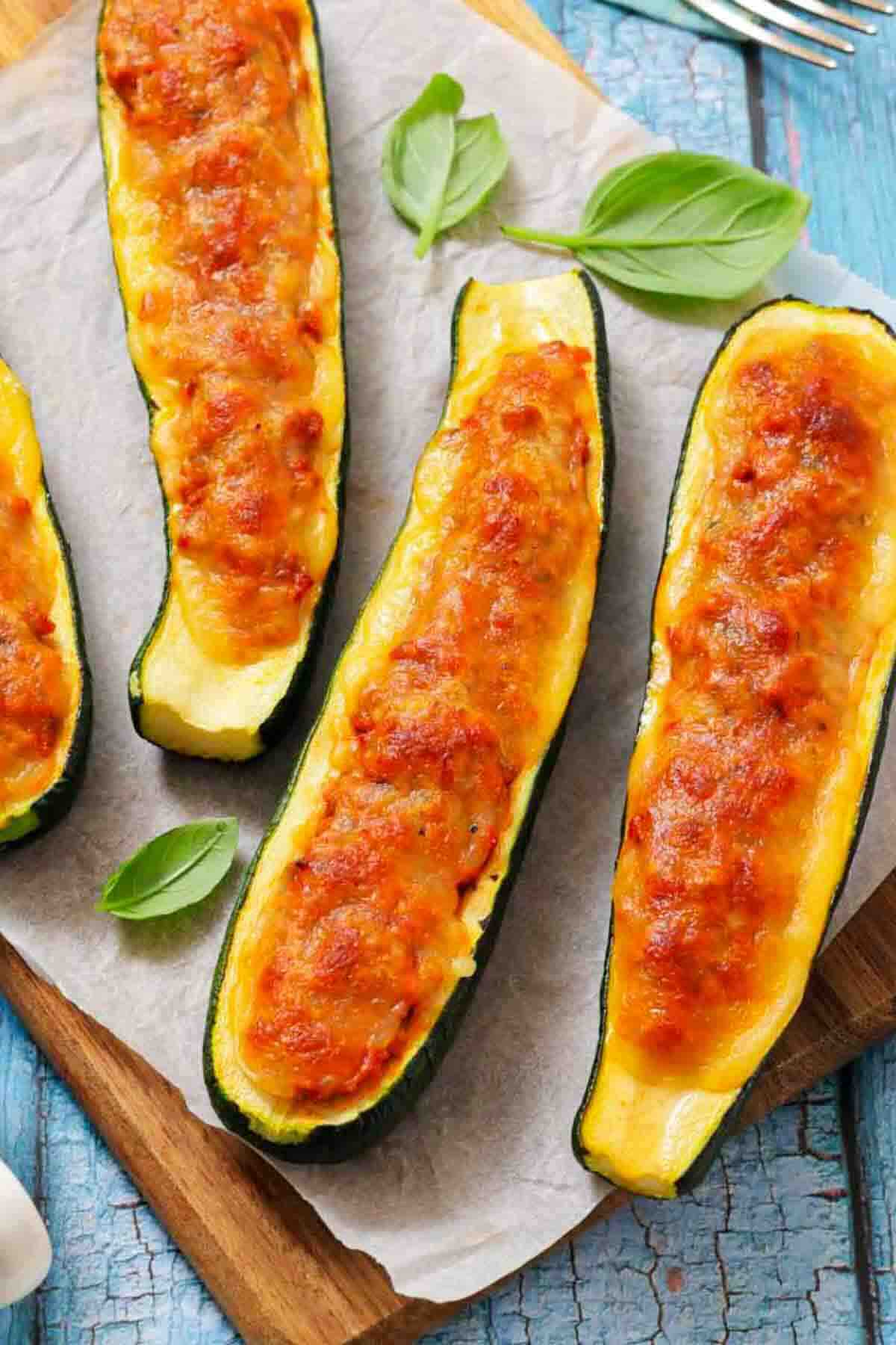 Four zucchini boats filled with a cheesy, golden-brown topping are arranged on parchment paper with fresh basil leaves on a wooden board.