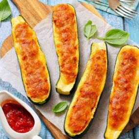 Four baked zucchini boats filled with melted cheese and tomato mixture, served on parchment paper with basil leaves and a side of tomato sauce.