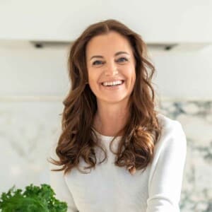 A woman with long brown hair wearing a white top smiles at the camera in a bright kitchen, with leafy greens visible in the foreground.
