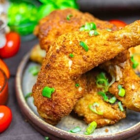 A close-up of crispy fried chicken drumsticks garnished with chopped green onions, served on a plate with tomatoes and greens in the background.