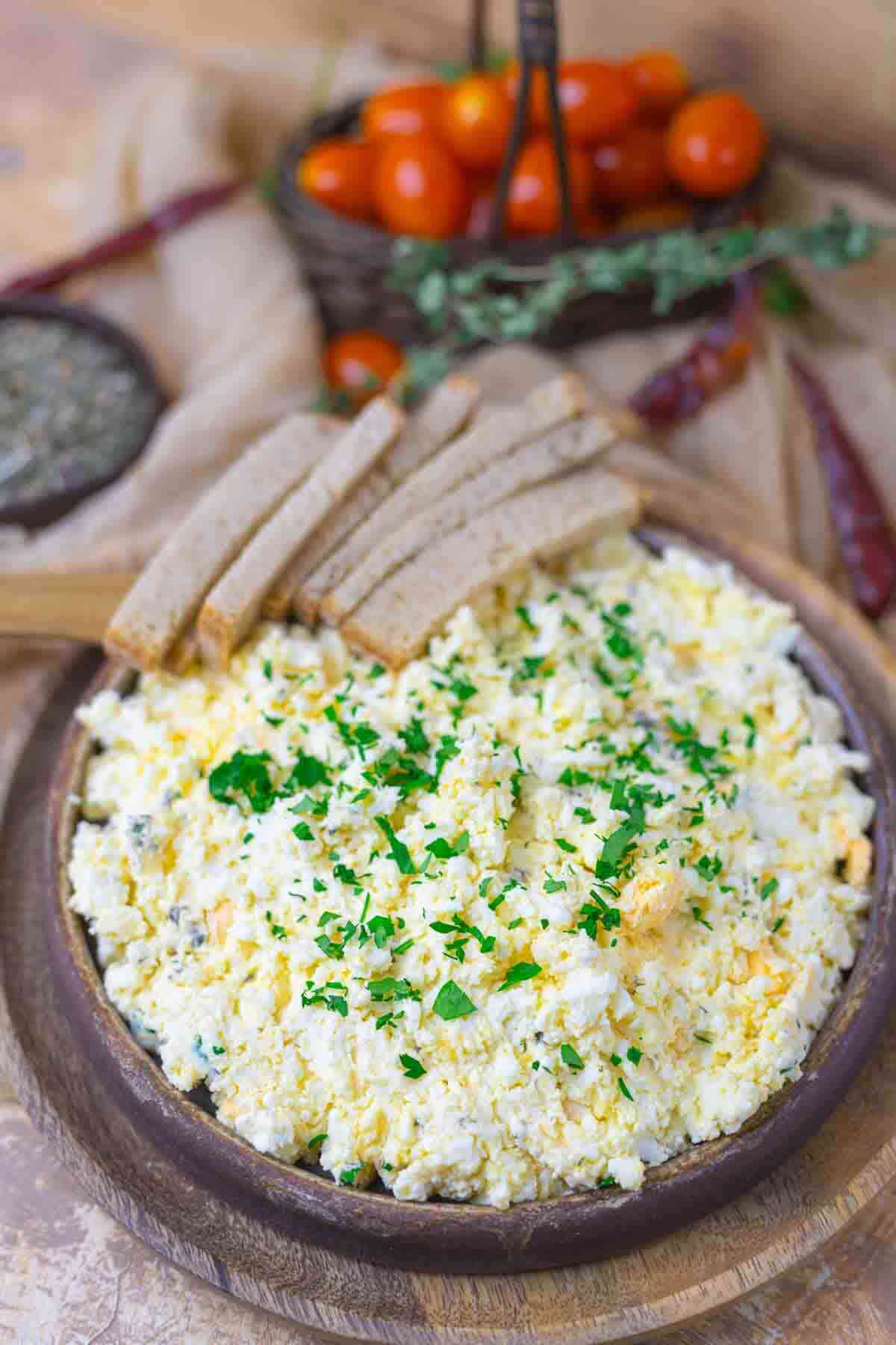 A wooden plate with a creamy egg salad garnished with chopped herbs, served with slices of bread; tomatoes and dried chilies are in the background.
