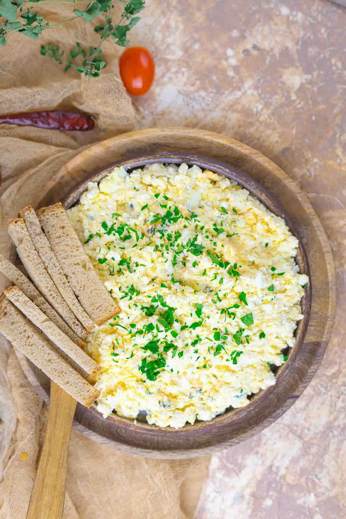 A wooden bowl of scrambled eggs garnished with chopped herbs, served with slices of brown bread on the side.