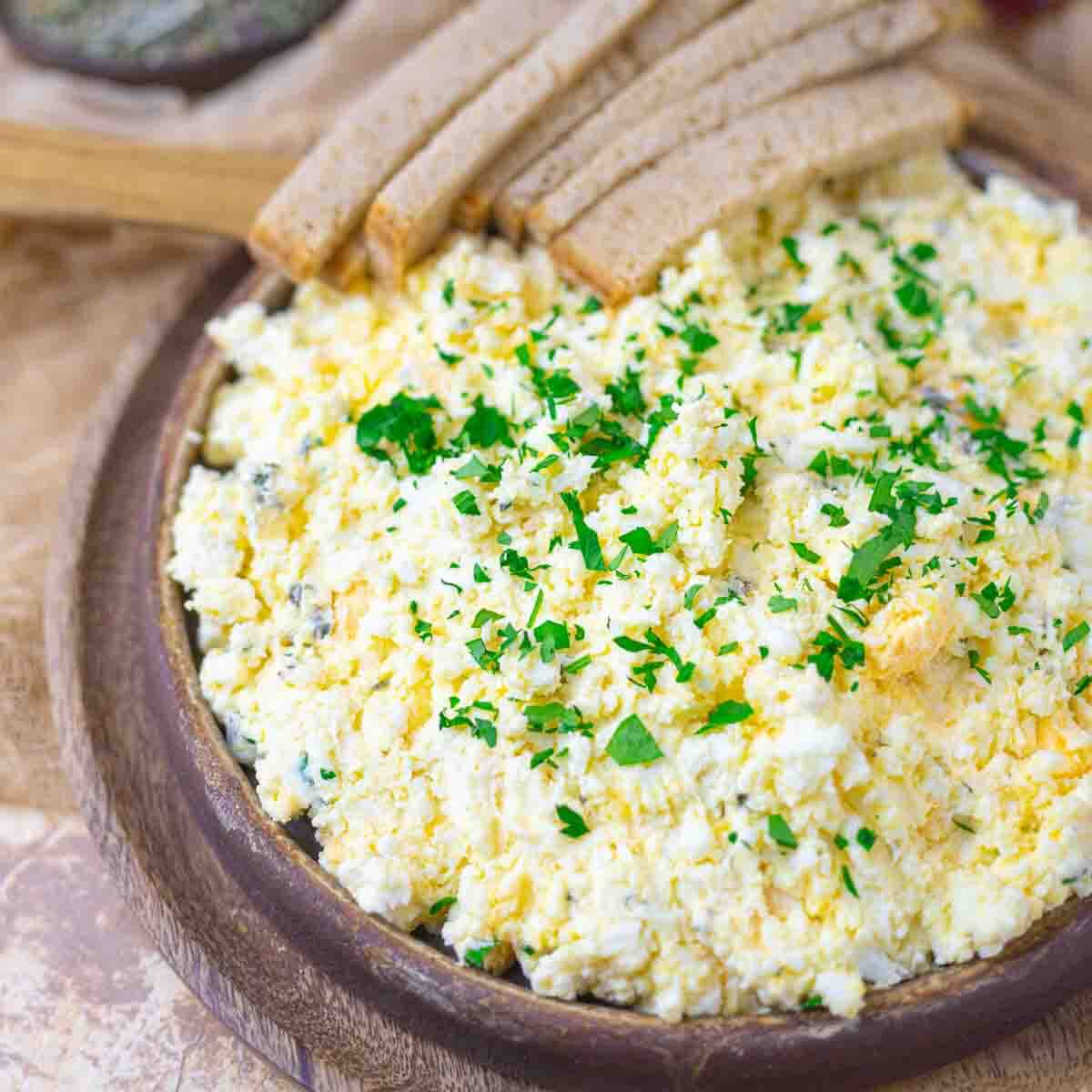 A wooden bowl filled with egg salad, garnished with chopped parsley, and served with several slices of brown bread on the side.