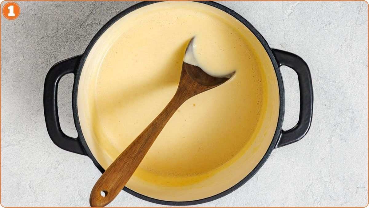A pot filled with creamy yellow sauce sits on a light surface, with a wooden spoon resting inside.