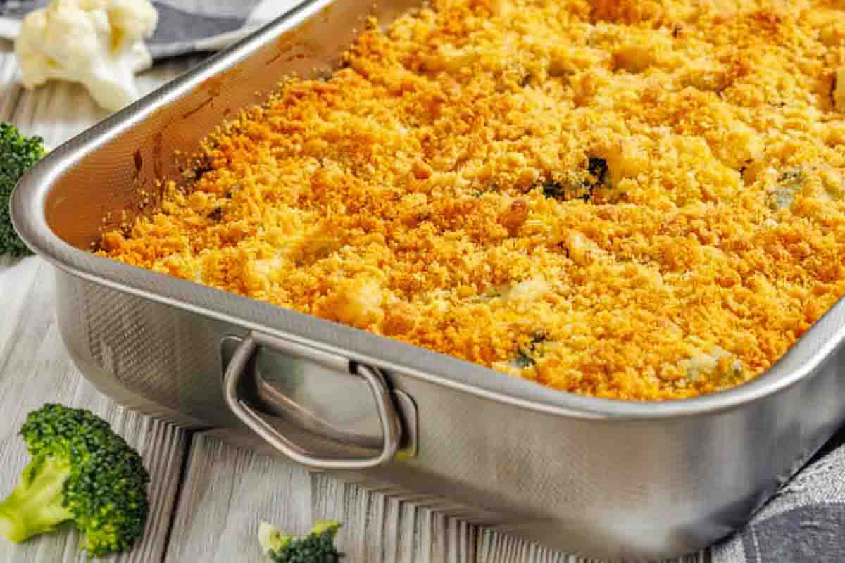 A metal baking dish filled with baked casserole topped with a golden breadcrumb crust, with pieces of broccoli nearby on a wooden surface.