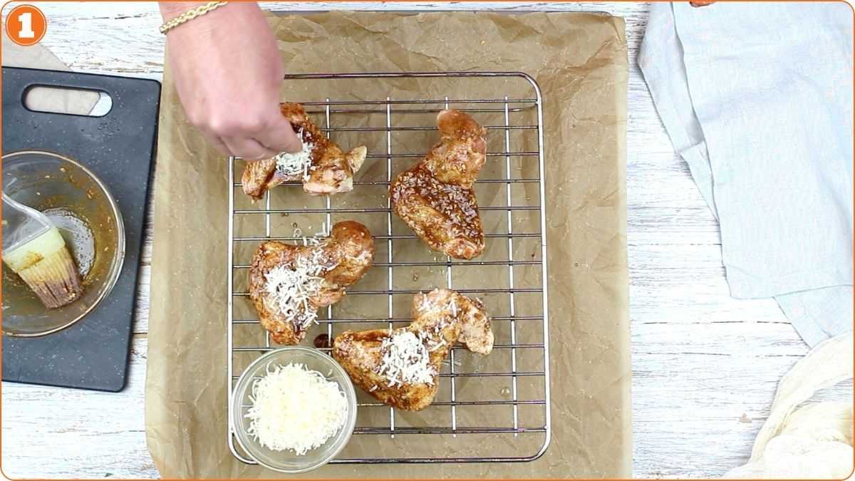 A hand sprinkles grated cheese onto seasoned chicken wings placed on a wire rack over a baking sheet, with a bowl of cheese and a basting brush nearby.