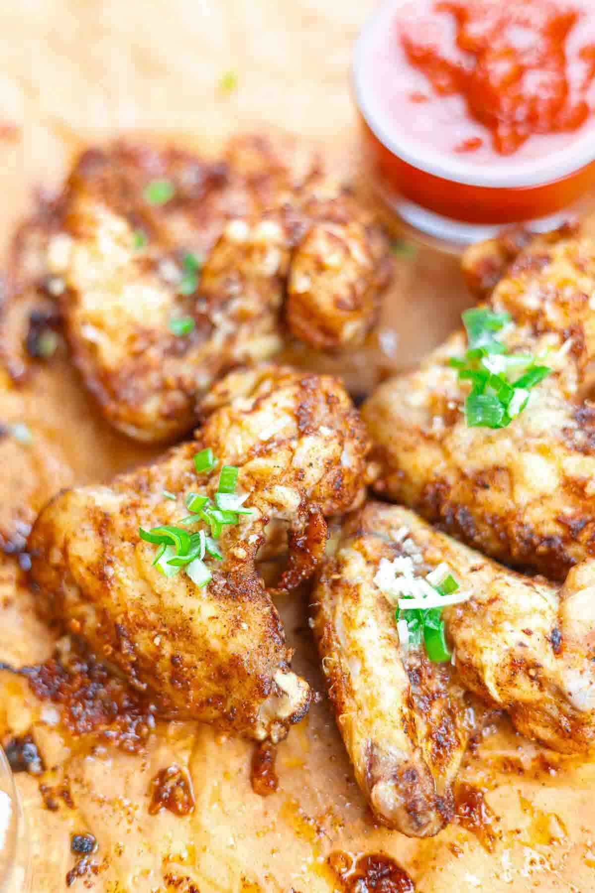 Close-up of several seasoned chicken wings garnished with chopped green herbs, served on parchment paper with a small cup of red dipping sauce.