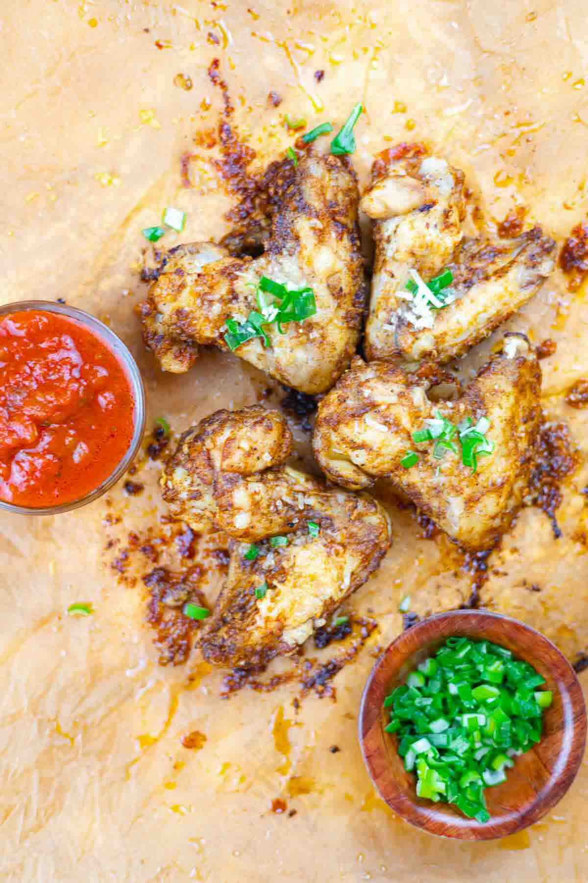 Five seasoned chicken wings on parchment paper, garnished with chopped green herbs, served with red sauce and a small bowl of chopped green onions.