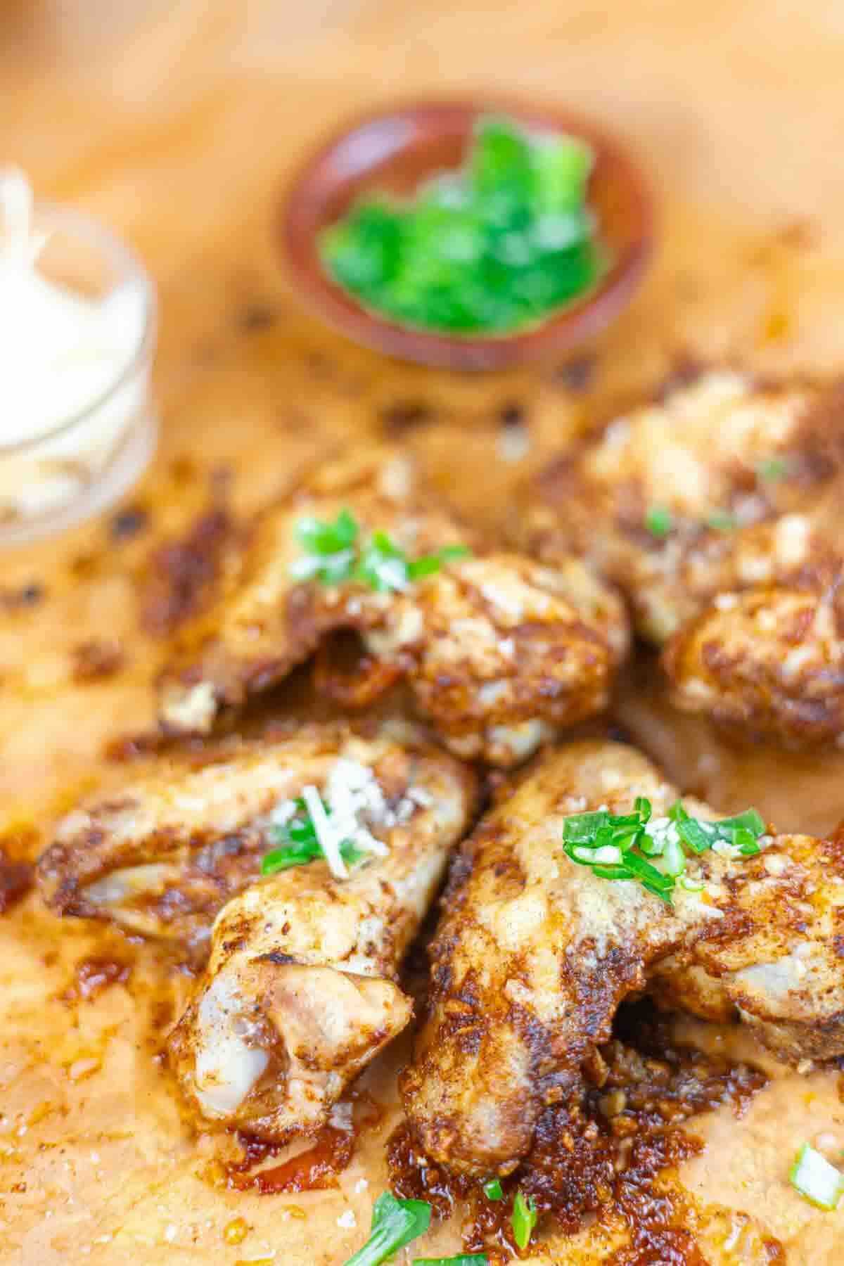 Close-up of seasoned and baked chicken wings garnished with chopped herbs, with small bowls of sauce and sliced onions in the background.