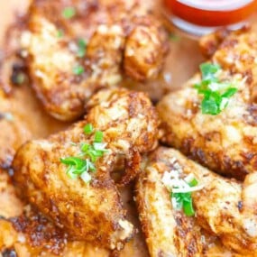 Close-up of seasoned and cooked chicken wings garnished with chopped green onions, with a small cup of red dipping sauce in the background.