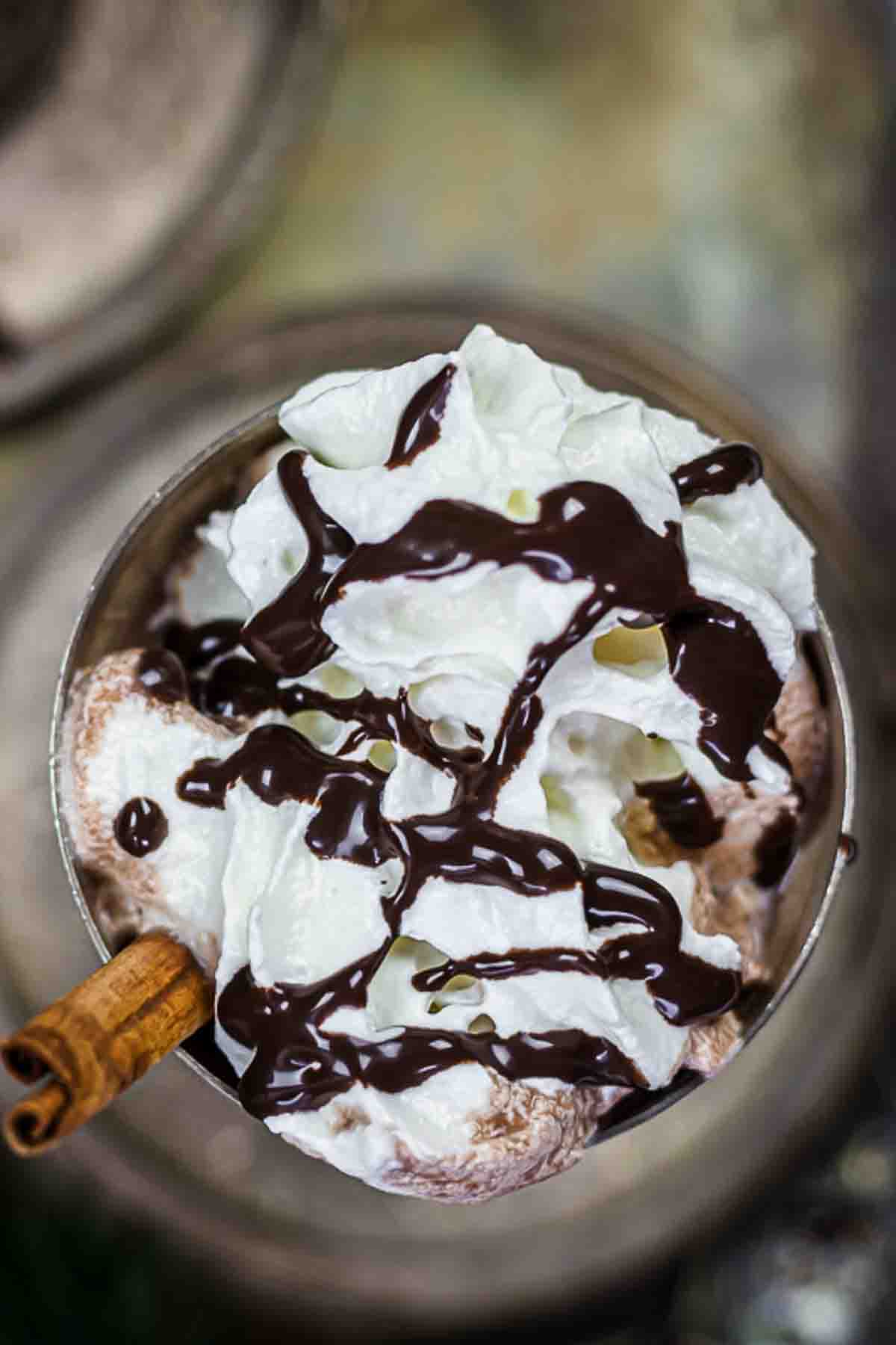 A close-up view of whipped cream topped with chocolate syrup in a glass, with a cinnamon stick on the side.