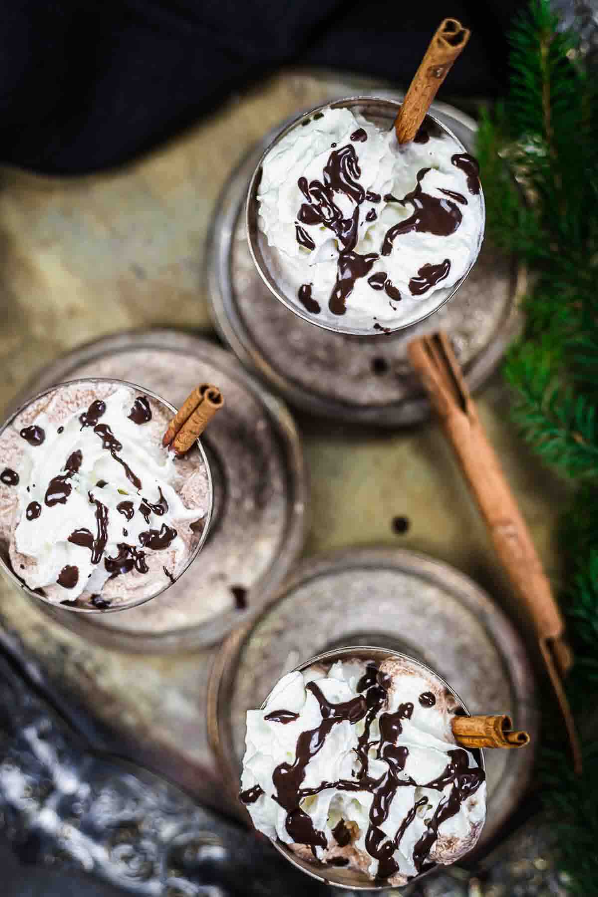 Three mugs of hot chocolate topped with whipped cream, drizzled with chocolate syrup, and garnished with cinnamon sticks, viewed from above.