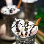 A silver cup filled with hot chocolate, topped with whipped cream, chocolate drizzle, and a cinnamon stick, with another similar cup blurred in the background.