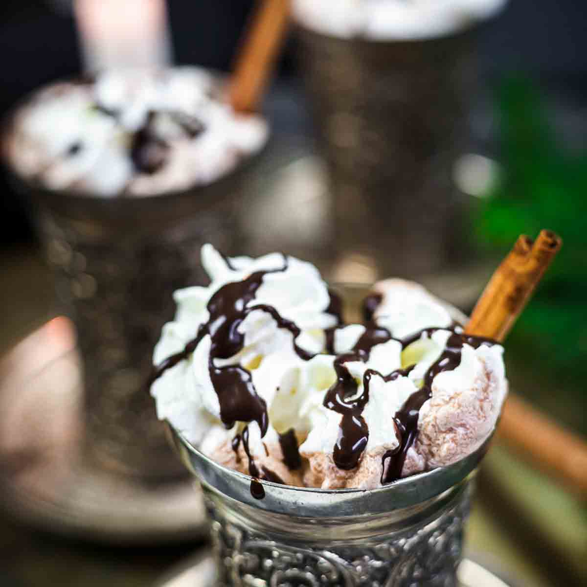 A silver cup filled with hot chocolate, topped with whipped cream, chocolate drizzle, and a cinnamon stick, with another similar cup blurred in the background.