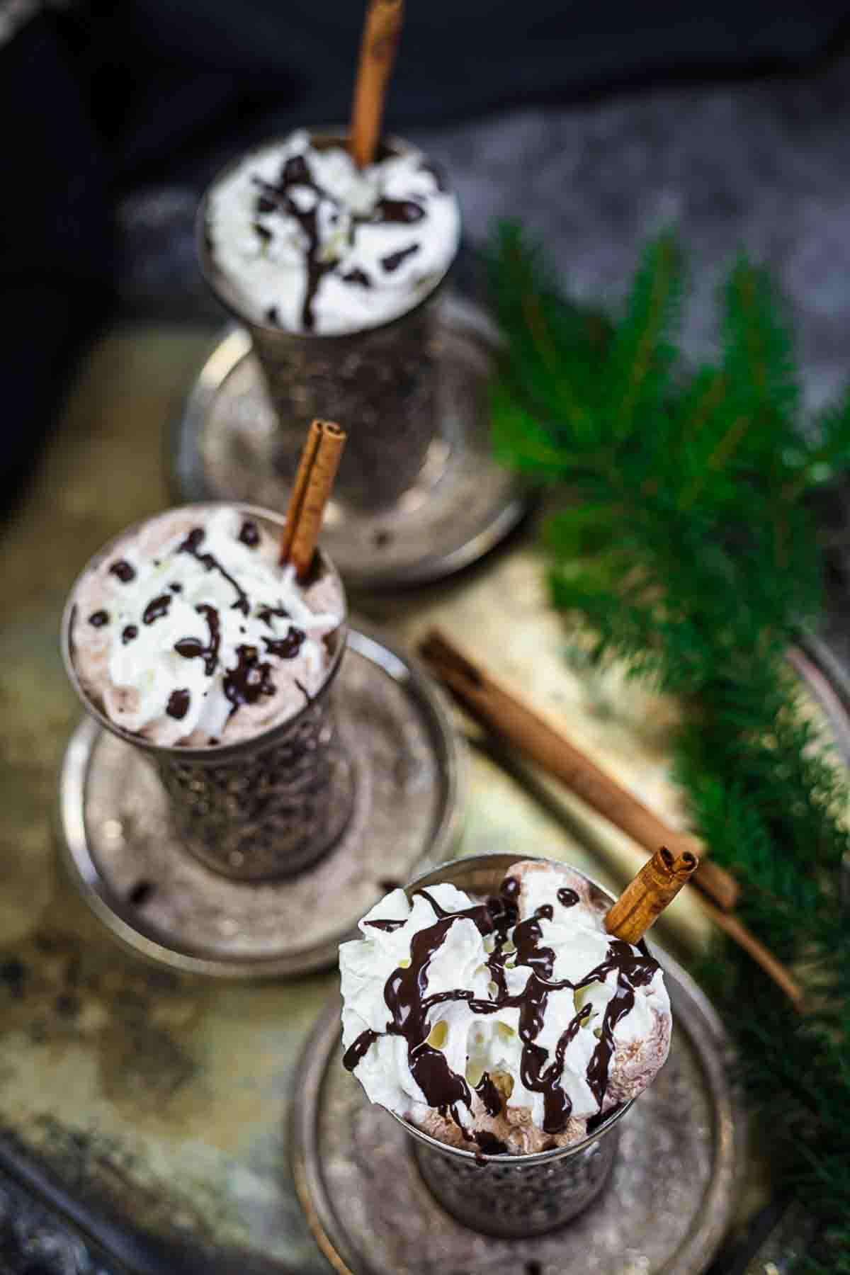 Three metal cups filled with whipped cream-topped hot chocolate, drizzled with chocolate syrup, and garnished with cinnamon sticks, placed on a tray with cinnamon sticks and greenery.
