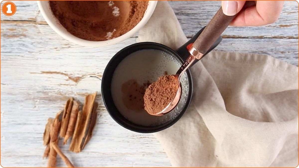 A hand holds a spoon of cocoa powder over a mug of milk on a wooden surface, with a bowl of cocoa powder and cinnamon sticks nearby.