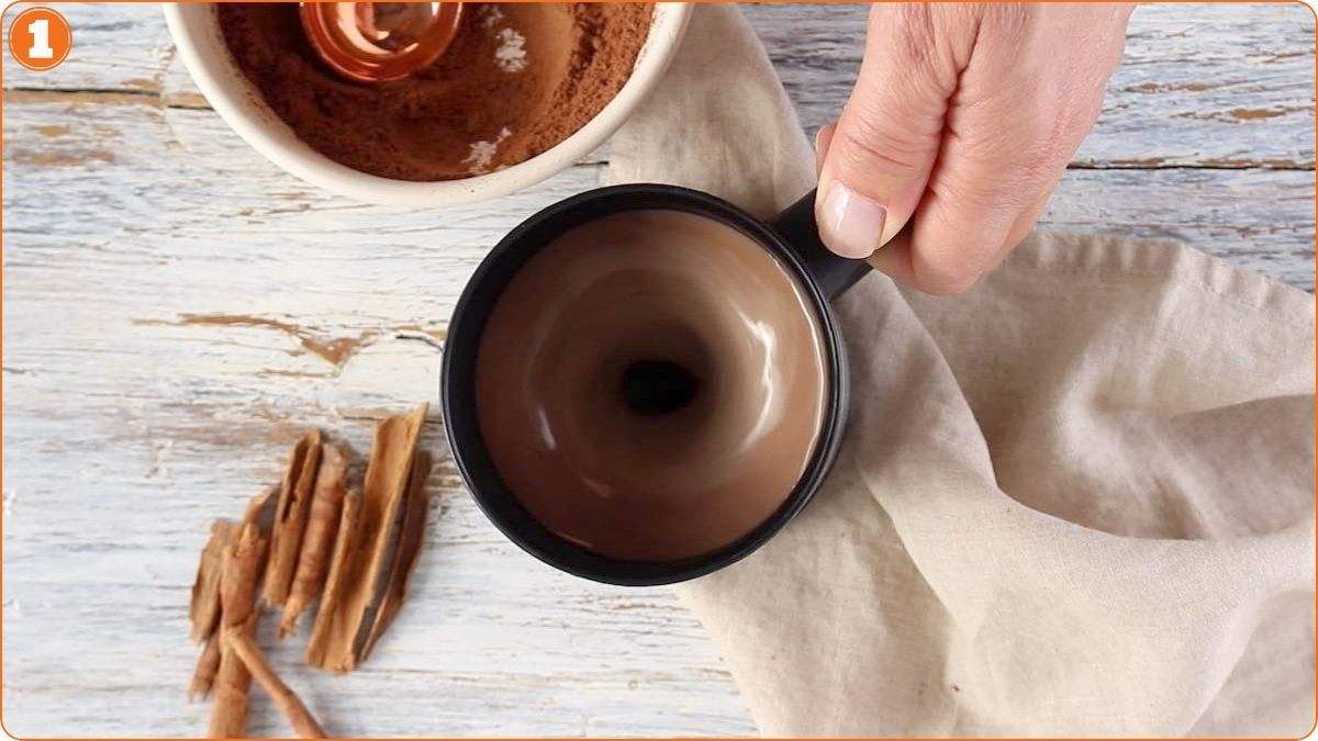 A hand holds a black mug of swirling hot chocolate next to a bowl of cocoa powder, cinnamon sticks, and a beige cloth on a rustic wooden surface.