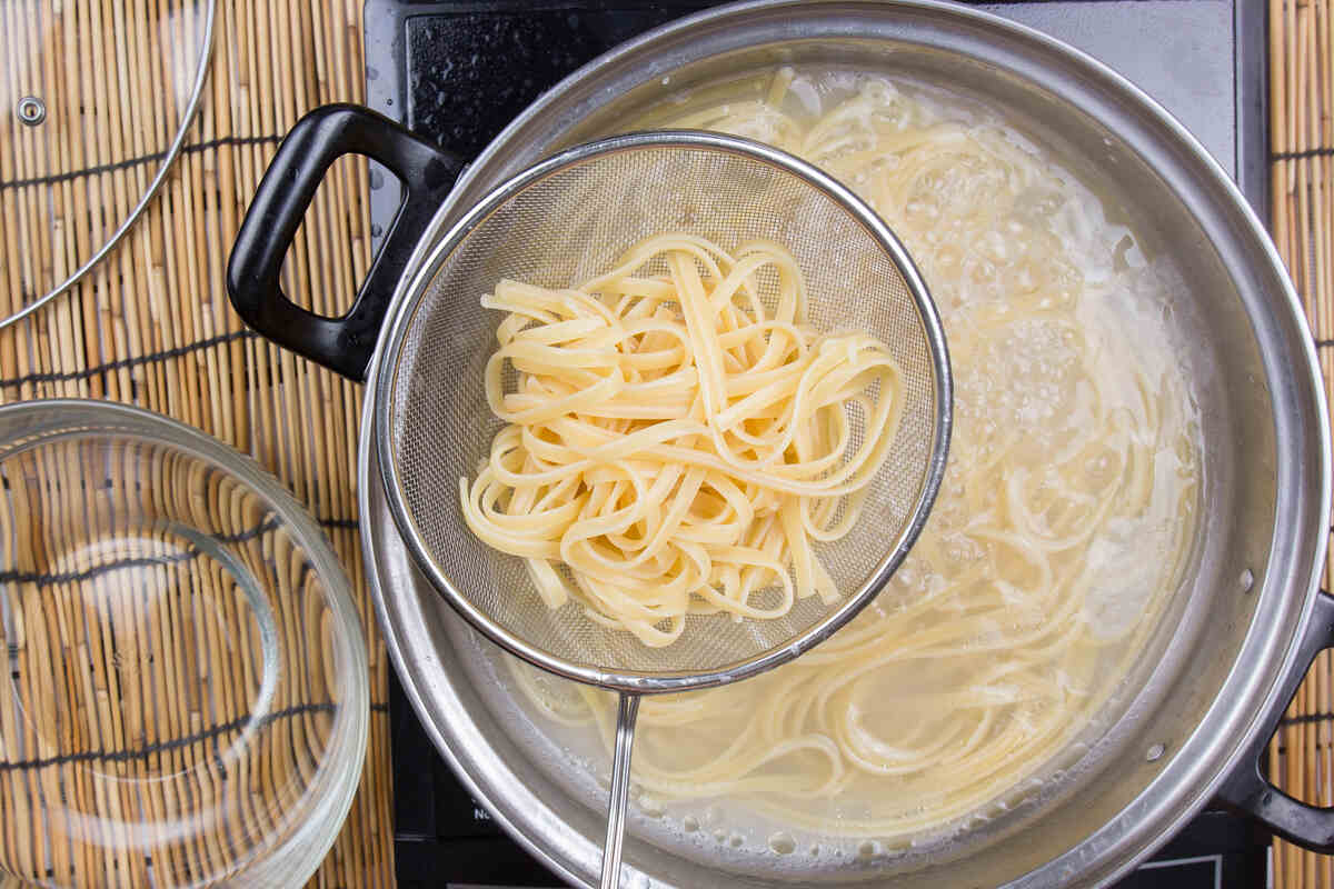 A strainer holds cooked linguine above a pot of boiling water on a stovetop, with more linguine visible in the pot and a glass bowl nearby.