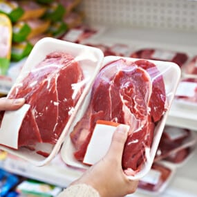 A person holds two packages of raw red meat in a grocery store meat section, comparing the products.