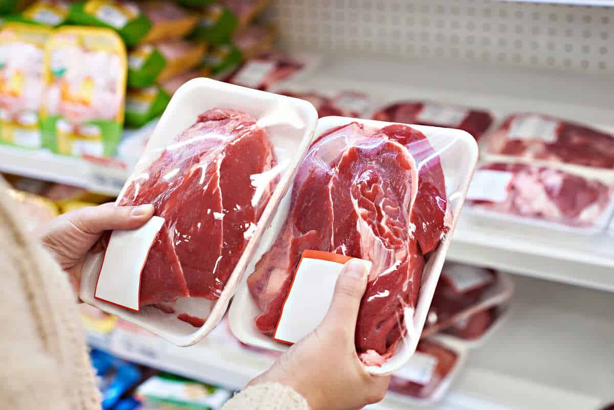 A person holds two packages of raw red meat in a grocery store meat section, comparing the products.