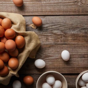 Brown eggs in a cloth-lined basket and white eggs in bowls are arranged on a wooden table, with a few eggs scattered around.