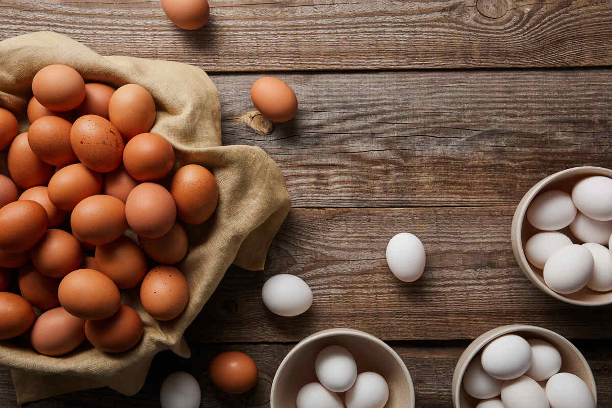 Brown eggs in a cloth-lined basket and white eggs in bowls are arranged on a wooden table, with a few eggs scattered around.