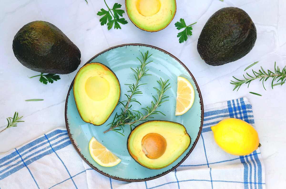 A plate with avocado halves, lemon slices, and rosemary, surrounded by whole avocados, a lemon, and herbs on a white cloth with a blue-checked towel.