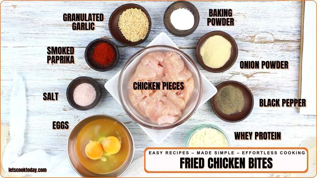 Top-down view of labeled fried chicken bites ingredients including chicken pieces, eggs, spices, whey protein, and baking powder arranged on a white wooden surface.